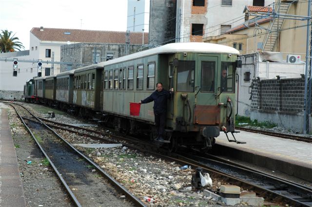 Maniobra a l'estació de Sàsser. Tren regular Sassari-Sorso composat per locomotora anys 50, 4 cotxes metàl-lics dels anys trenta i un furgó correu de fusta més antic. Recorregut: 11 kms.
<br />Nota: el banderí és verd per l'altra banda.